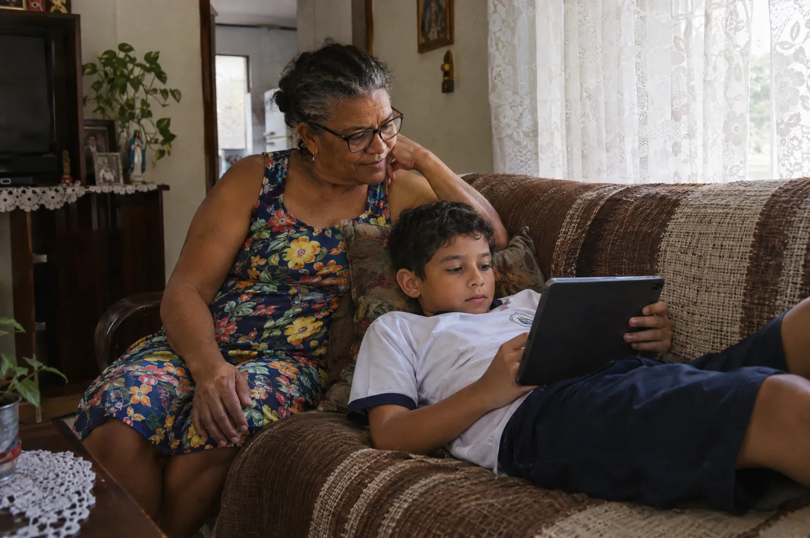 Avó acompanha o neto, que faz uma atividade da escola no tablet, no sofá de casa