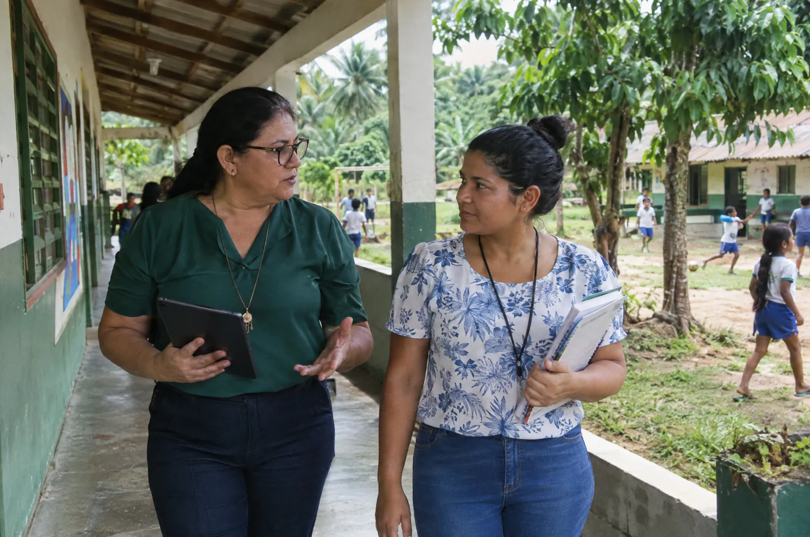 Diretora conversa com uma professora no corredor da escola, segurando um tablet