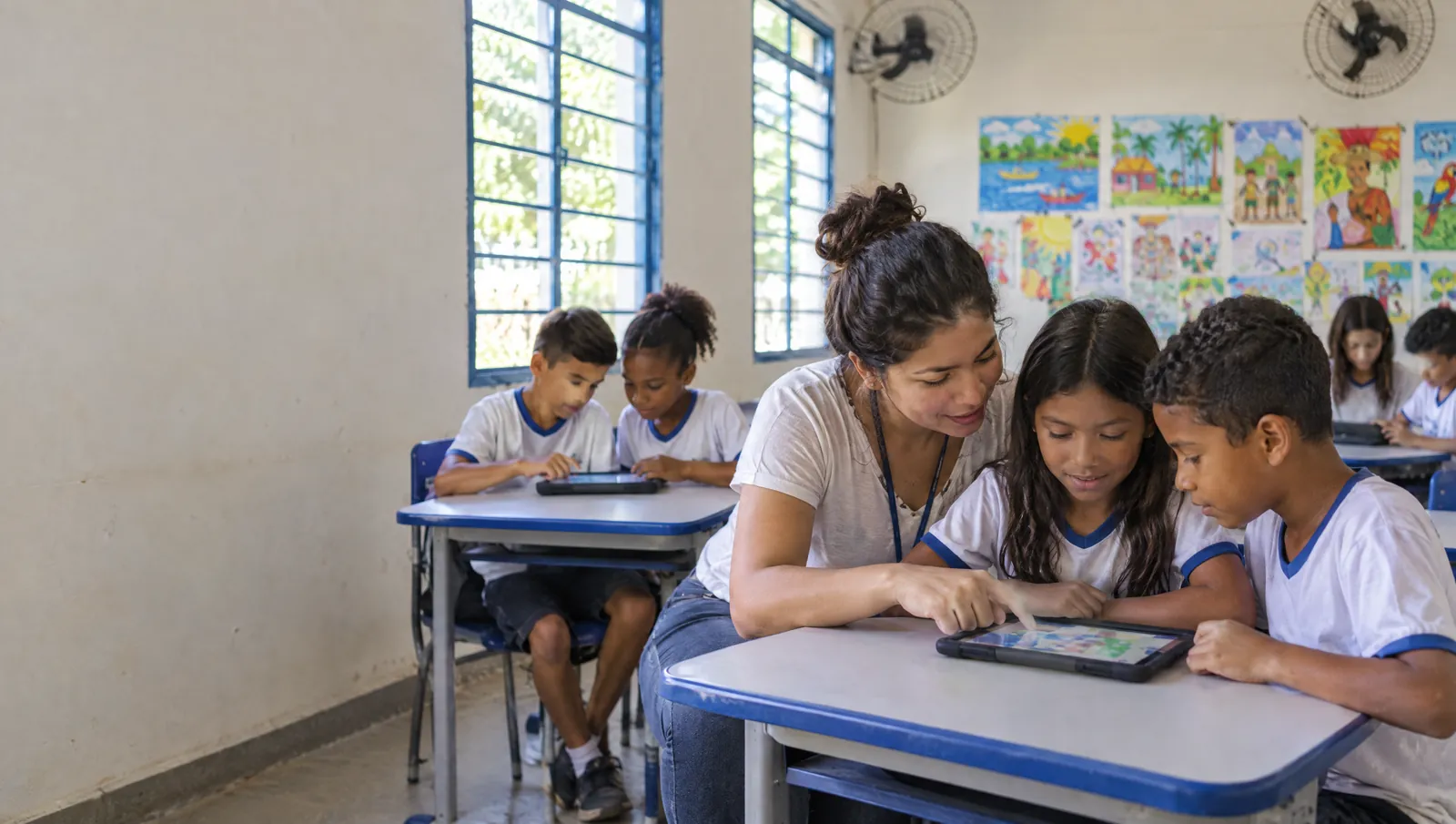 Professora ajuda dois alunos com um tablet numa sala de aula de escola pública